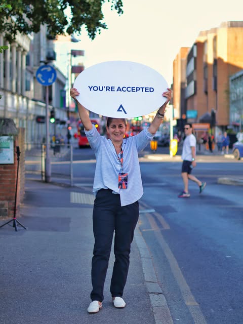 A woman smiles on a London street, holding up a sign that reads 'You're accepted'